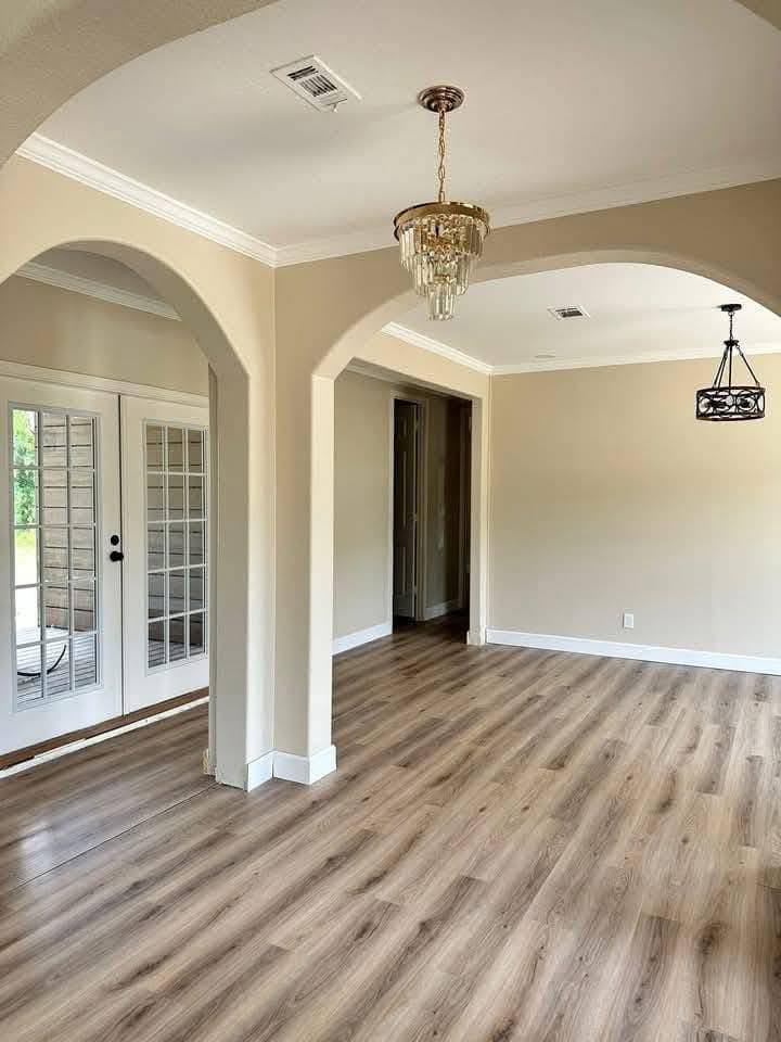 Elegant, empty room with beige walls and light wood flooring. Features arched doorways, a crystal chandelier, and French doors leading outside.