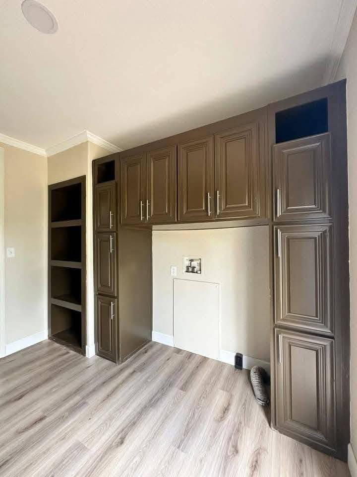 Empty laundry room with brown cabinets and light wood flooring. The wall has connections for a washing machine. The space feels tidy and organized.