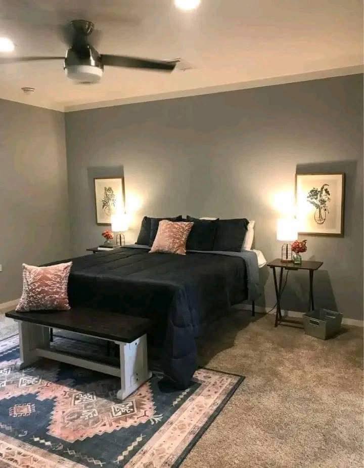 A cozy bedroom with a double bed features dark bedding and patterned pillows. Two bedside tables hold lamps and flower vases. Art decorates gray walls.