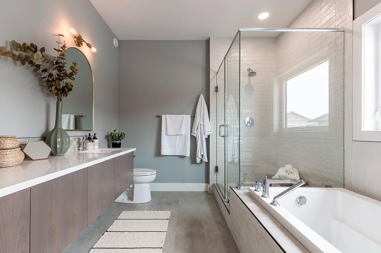 Modern bathroom with grey walls, a floating vanity, and a large mirror. Glass shower and white bathtub on the right. Bright and minimalist design.
