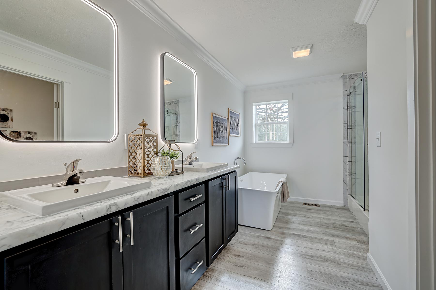 Modern bathroom with dual sinks, sleek black cabinetry, marble countertop, and illuminated mirrors. Features a freestanding tub and wood-like flooring.