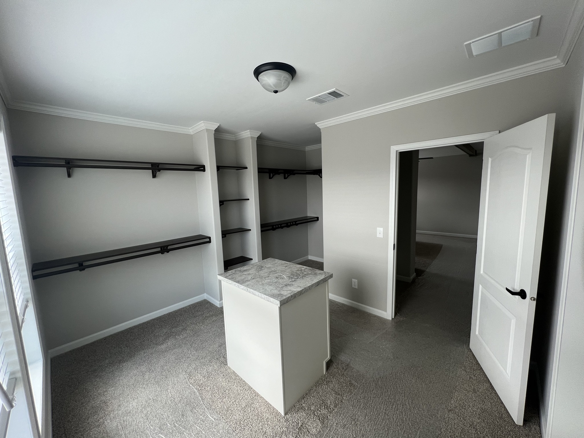 A spacious, well-lit walk-in closet with beige walls and carpet features dark wooden shelves and a marble-topped island. An open door leads to an adjacent room.