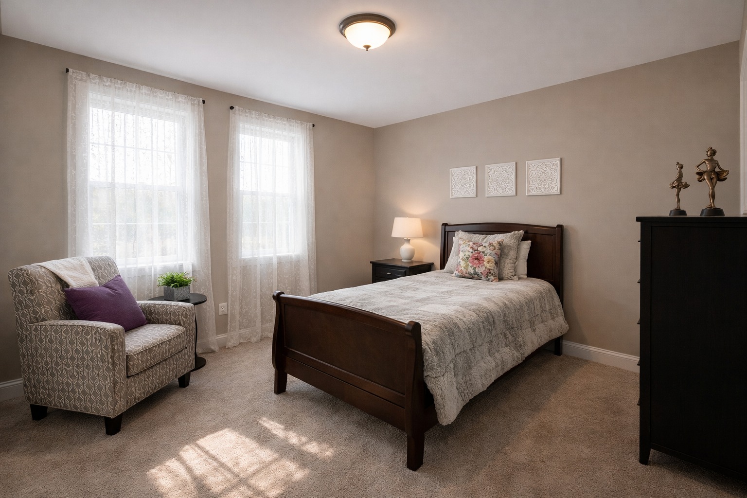 A cozy bedroom with beige walls features a wooden bed with floral bedding, a patterned armchair, and soft lighting. Two windows with sheer curtains let in natural light.