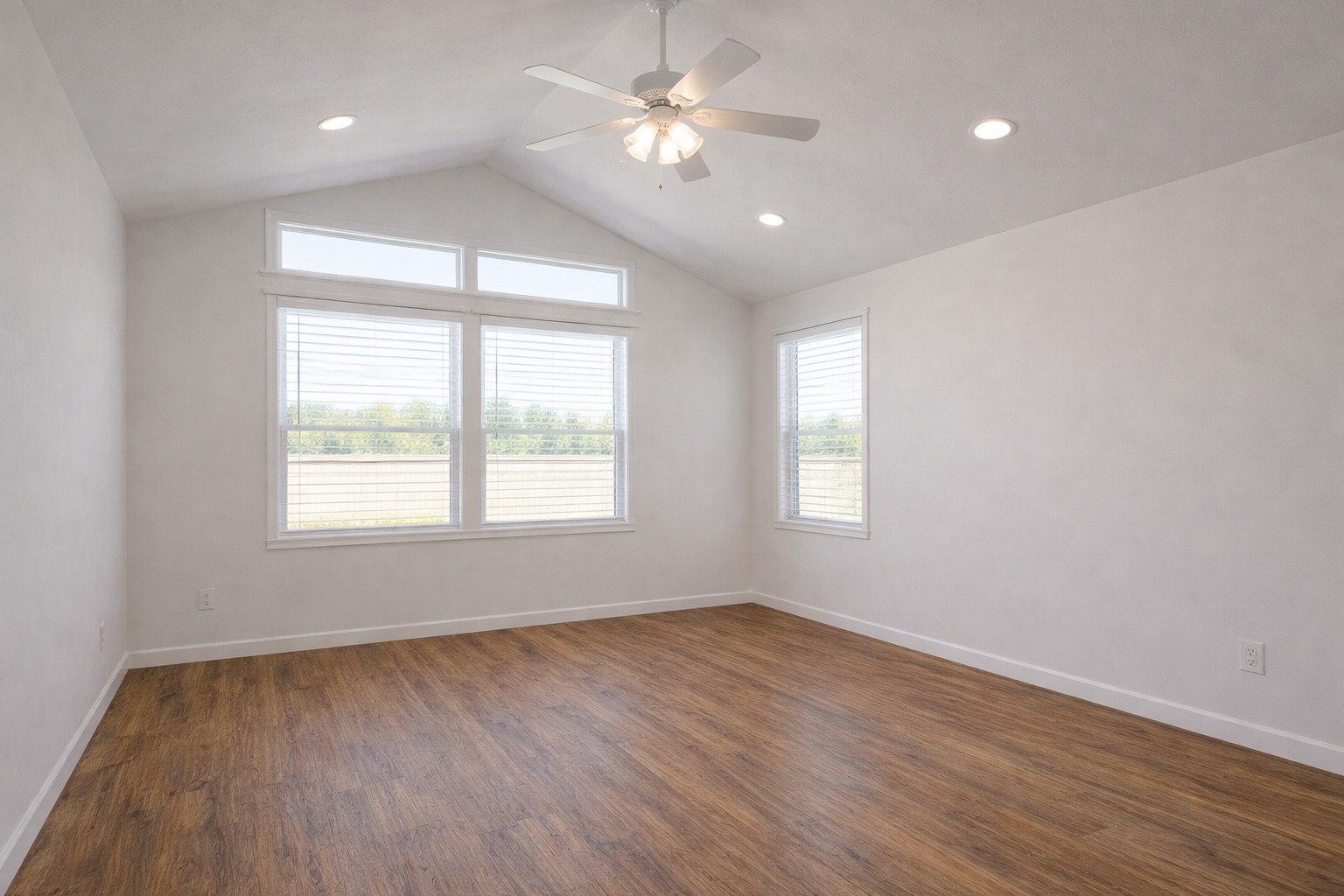 Bright, empty room with a vaulted ceiling, wooden floor, ceiling fan, and large windows. Natural light creates a welcoming, airy atmosphere.