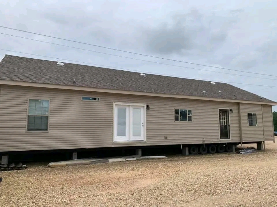 Single-story beige mobile home with a gabled roof, set on a gravel lot. It features windows, a double glass door, and a porch light under a cloudy sky.