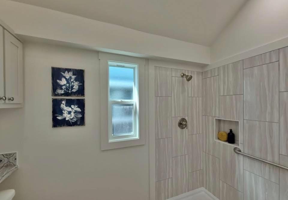 A modern bathroom with light wood paneling on the shower walls, a small window, and two wall art pieces with white floral designs on a dark background.