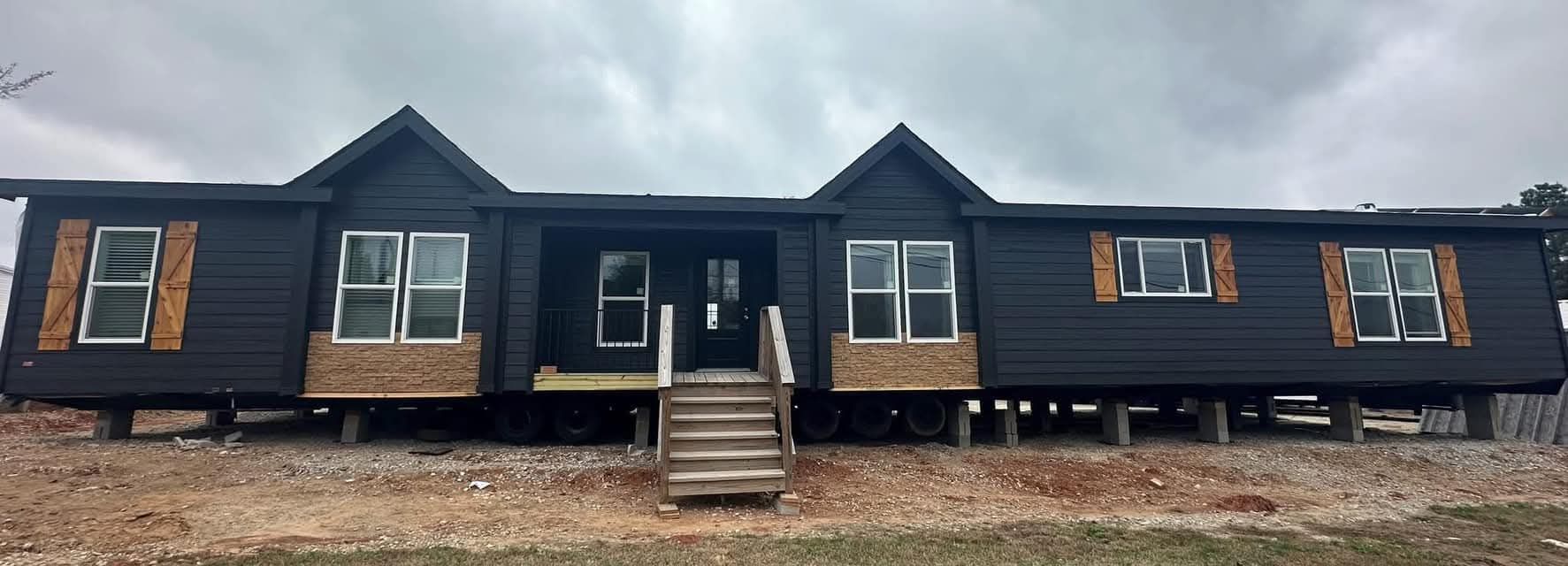 A large, dark gray modular home elevated on wheels, featuring multiple windows with wooden shutters and a central front door. The sky is overcast.