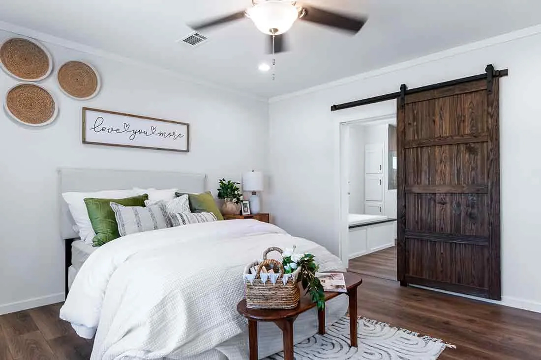 Cozy bedroom with a white bed, green and striped pillows, and a rustic sliding barn door. A small table holds a basket. Wooden floor adds warmth.