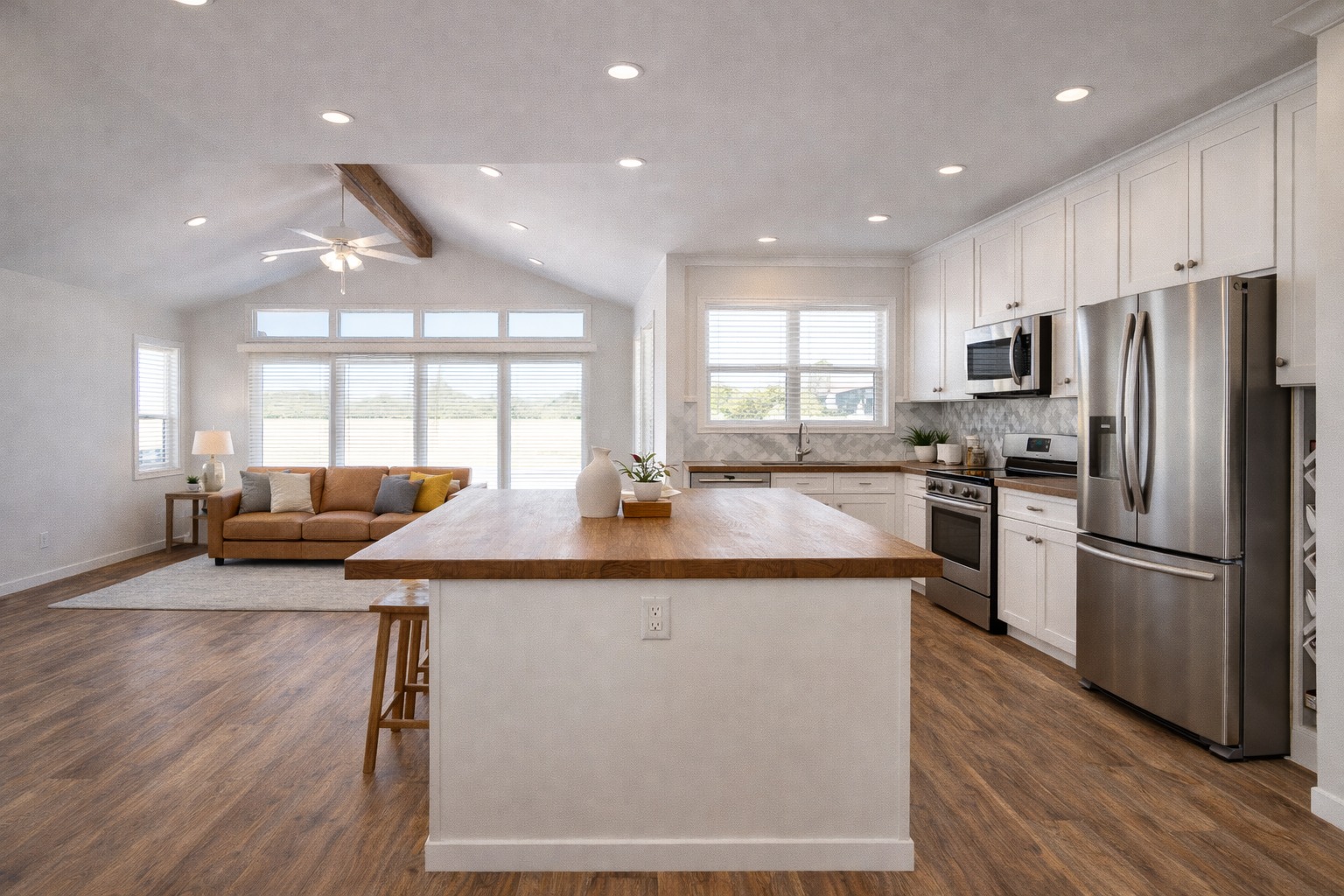 Spacious open-concept kitchen with wooden floors, white cabinets, stainless steel appliances, and a central island. Cozy sofa area with bright windows.