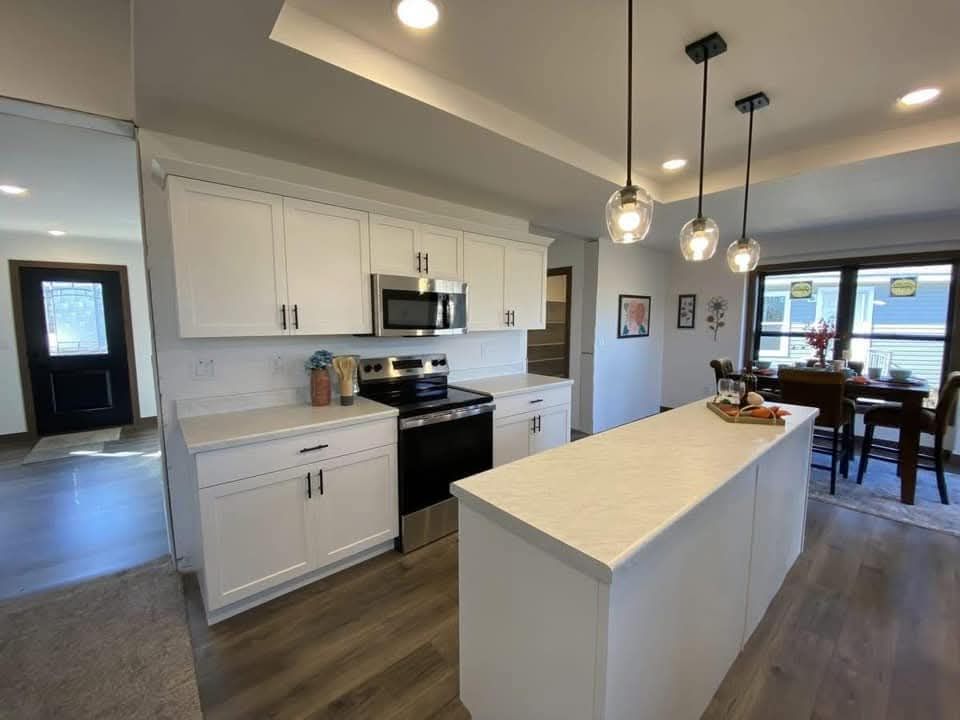 Modern kitchen with white cabinets, stainless steel appliances, and a sleek island under pendant lights. Dining area with large windows in the background.