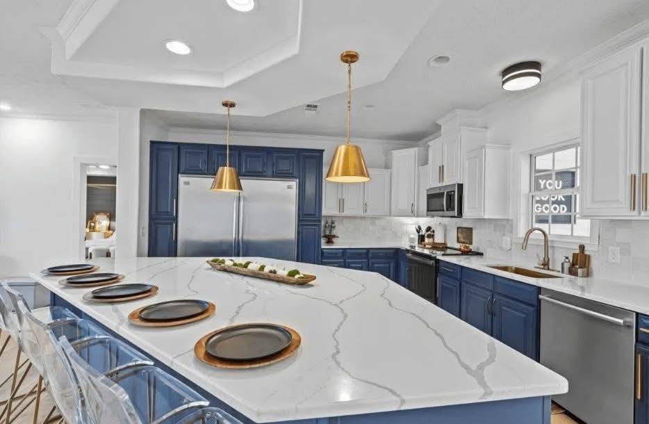 Spacious modern kitchen with white marble island, blue cabinetry, gold pendant lights, and clear chairs. Warm, inviting ambiance with sleek appliances.