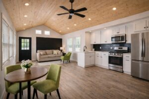 Modern open-plan living space with a wood ceiling, black ceiling fan, and warm wood flooring. Features a cozy kitchen, beige sofa, and green chairs by a round table.