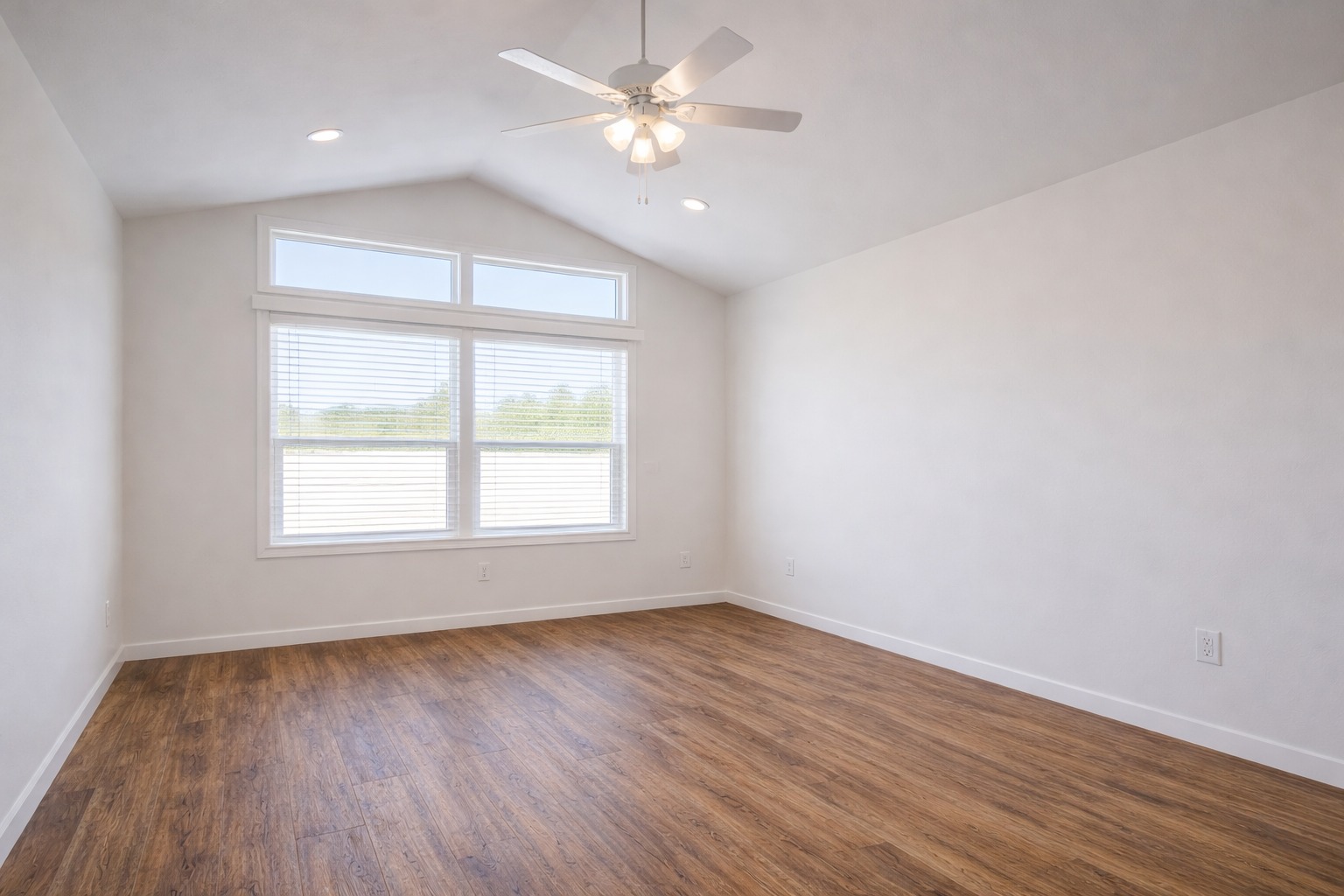 Empty room with vaulted ceiling, ceiling fan, large window with blinds, and hardwood floor. Bright and airy with natural light and a minimalist feel.