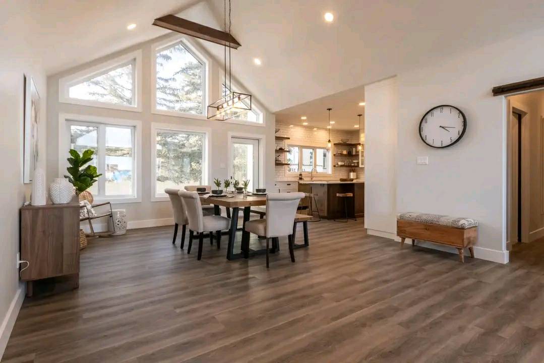 Spacious modern dining room with high ceiling, large windows, wooden table, white chairs, a geometric chandelier, and adjacent kitchen. Warm tones.