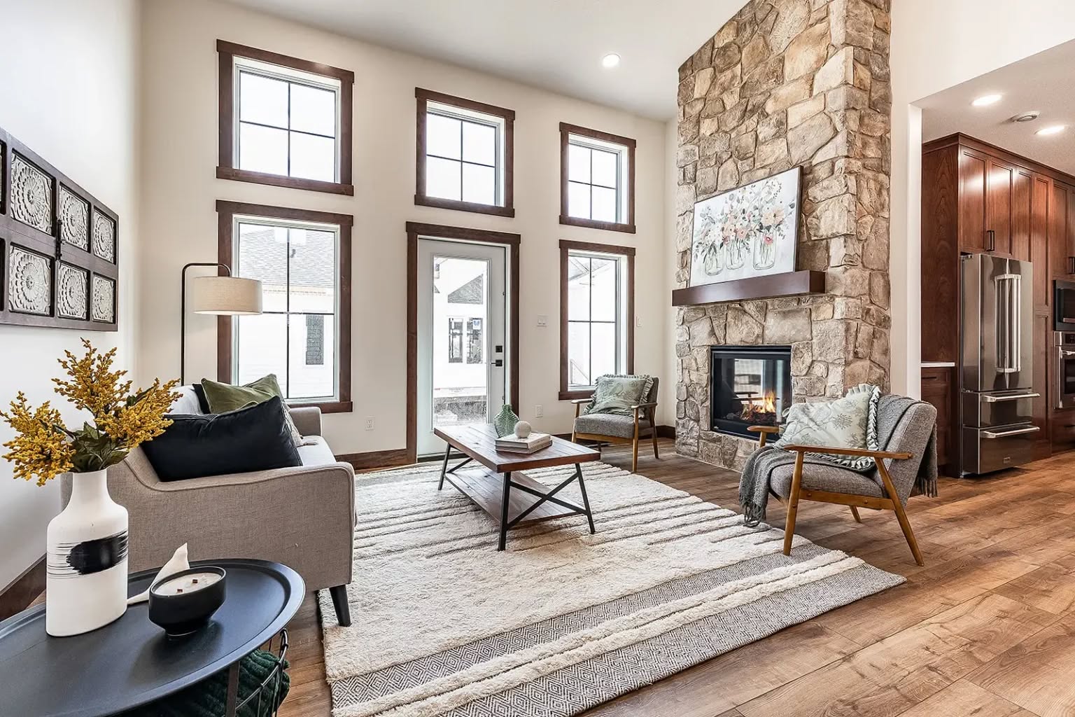 Warm living room with a cozy stone fireplace, modern furniture, and high ceilings. Large windows and natural light create an inviting atmosphere.