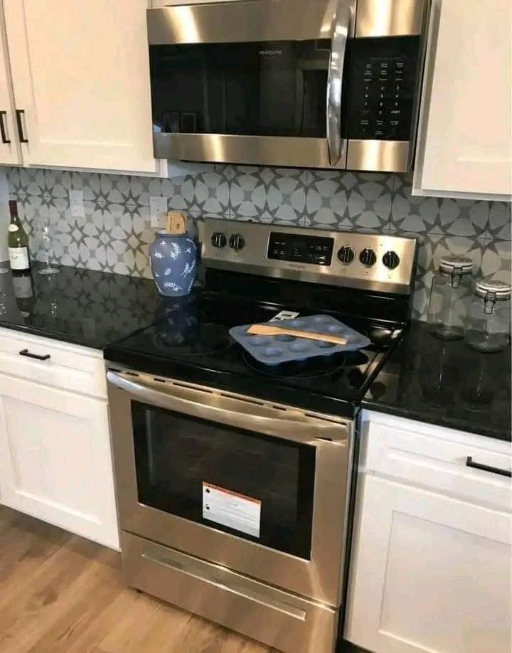 Modern kitchen scene with stainless steel stove and microwave, geometric tile backsplash, and white cabinets. A muffin tray rests on the stovetop.