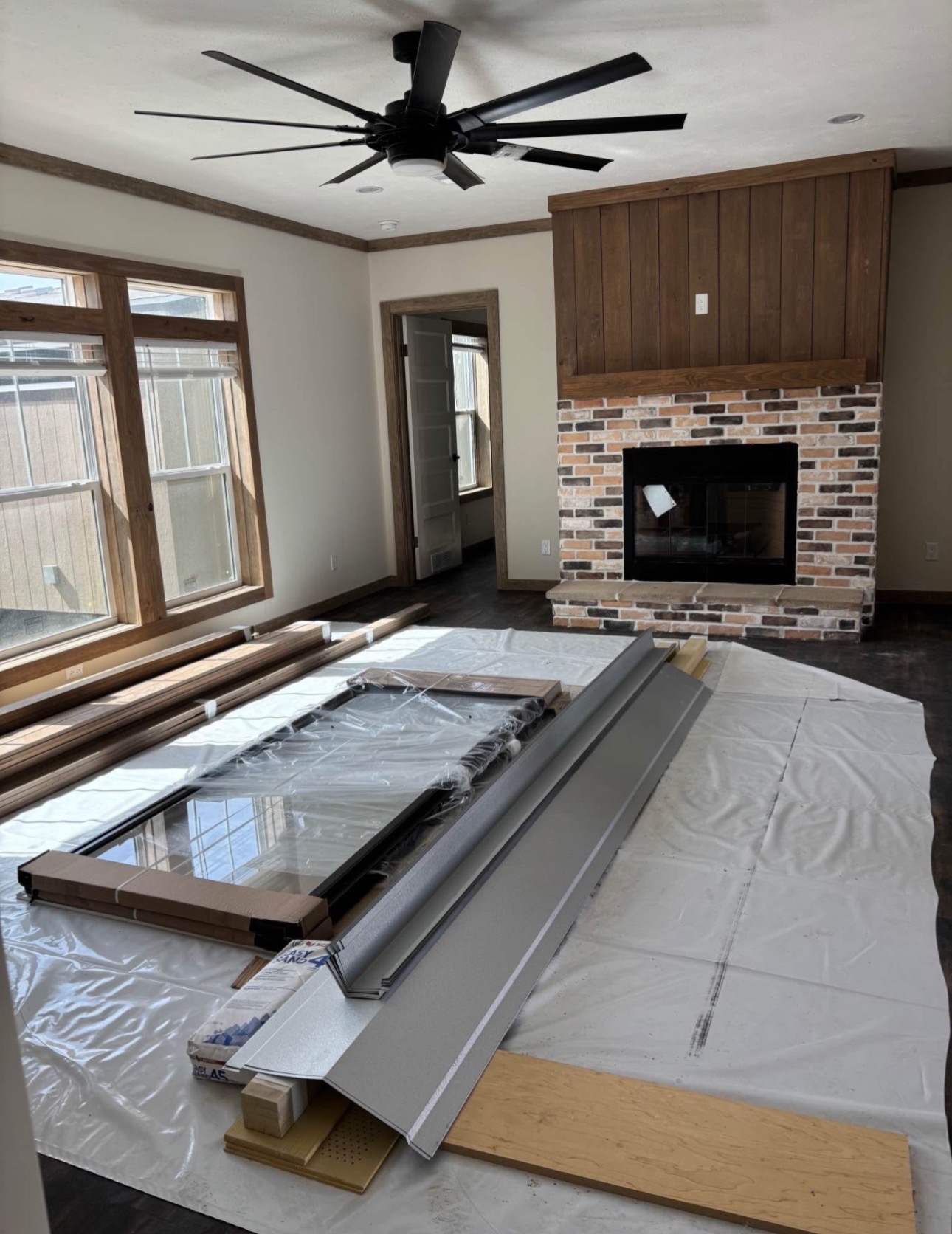 Living room under renovation with large windows, a modern ceiling fan, and a brick fireplace. Construction materials are laid on the floor.