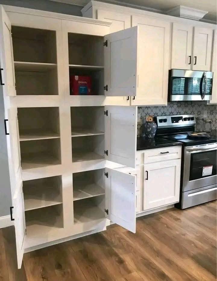 Open kitchen cabinet with eight empty shelves, except for a small red and blue item on the top shelf. Nearby stove and microwave, on wooden floor.