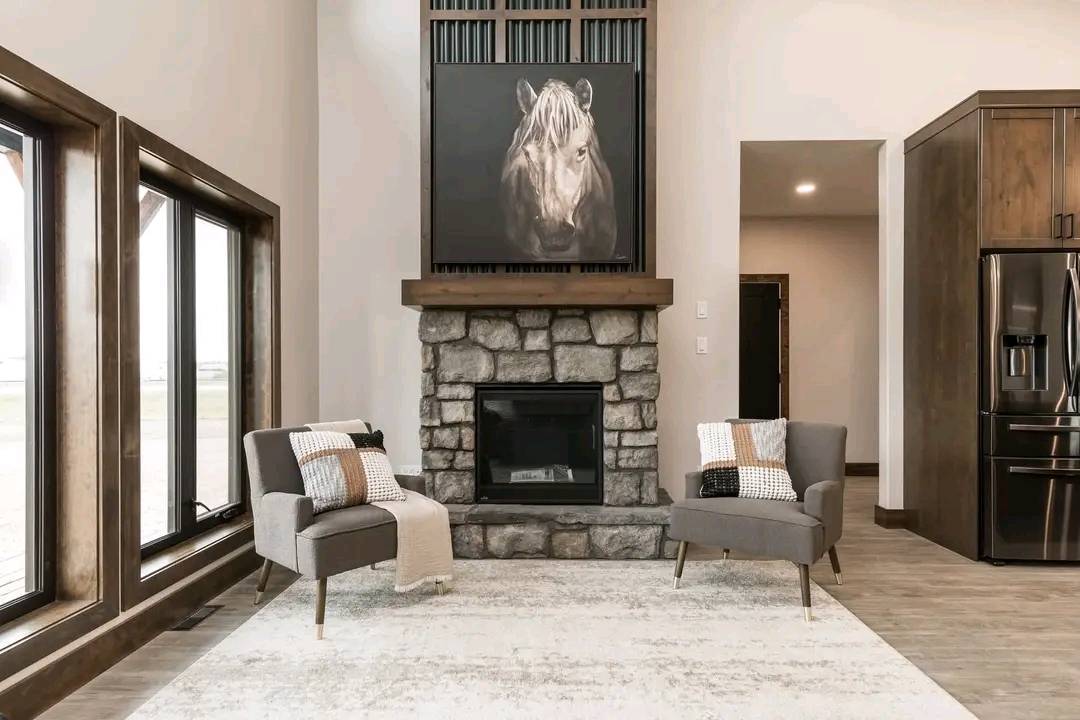 Modern living room with a stone fireplace, horse artwork, and two gray chairs. Sunlight streams through large windows, creating a serene atmosphere.