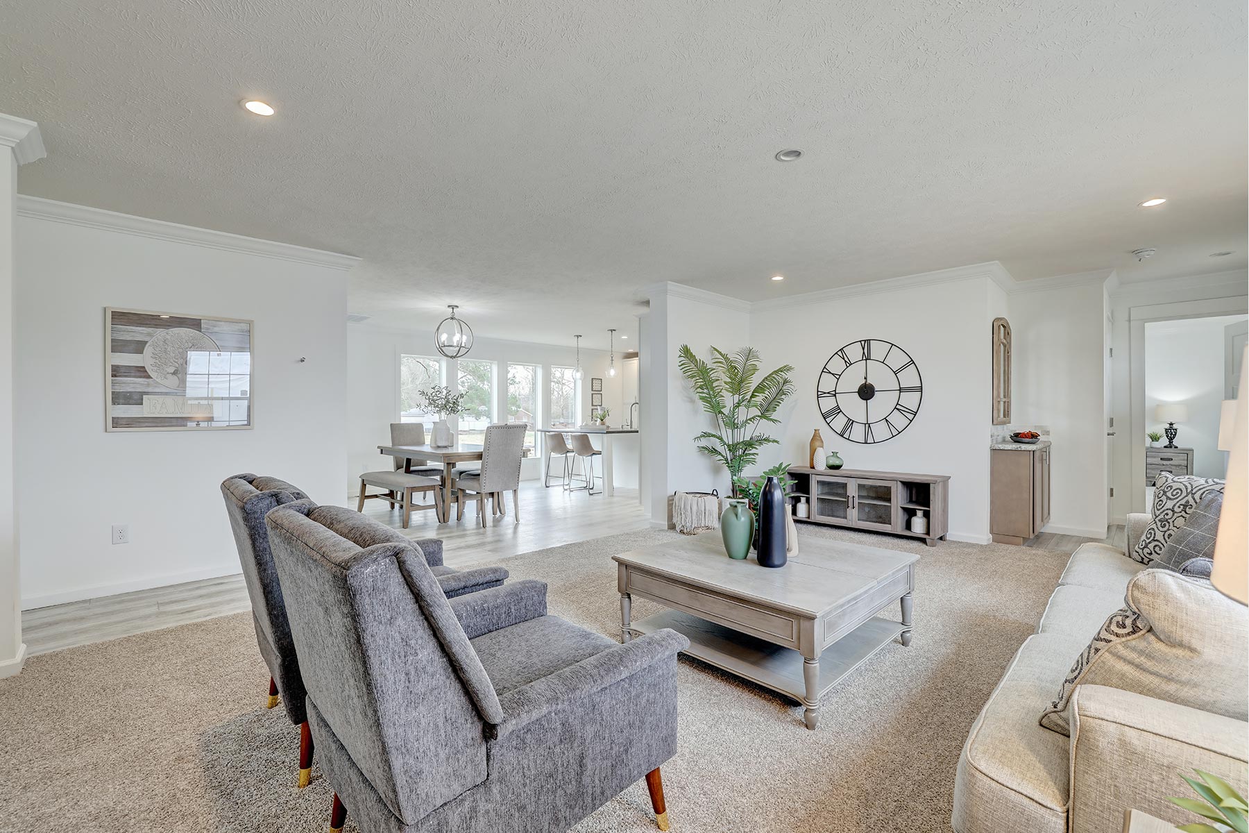 Spacious, modern living room with gray armchairs, a beige sofa, and a wooden coffee table. Large clock on wall, dining area and kitchen visible.