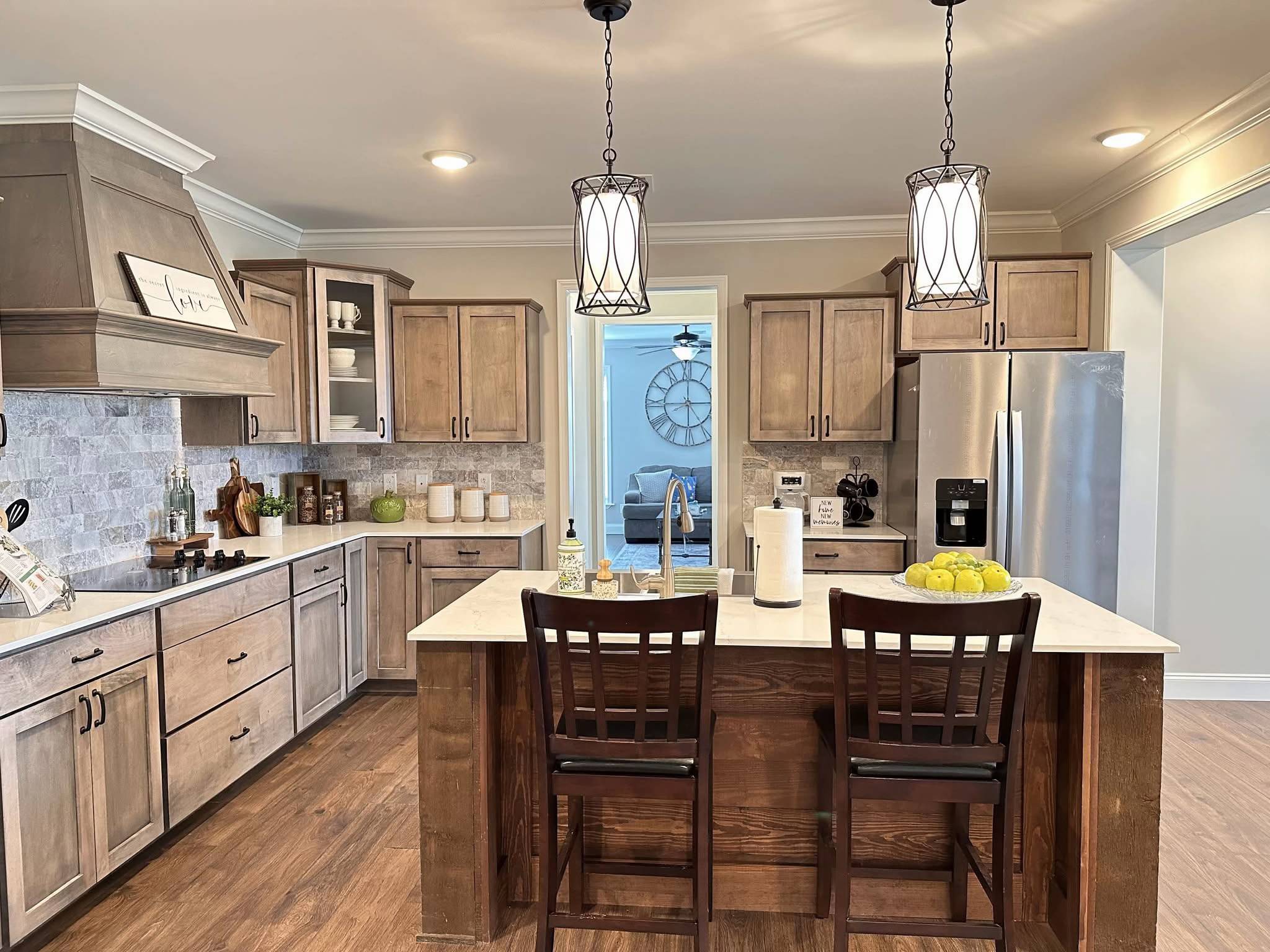 Bright kitchen with wooden cabinets, marble countertops, and two pendant lights. A central island with stools, fruit bowl, and a glimpse of a living room. Warm and inviting atmosphere.