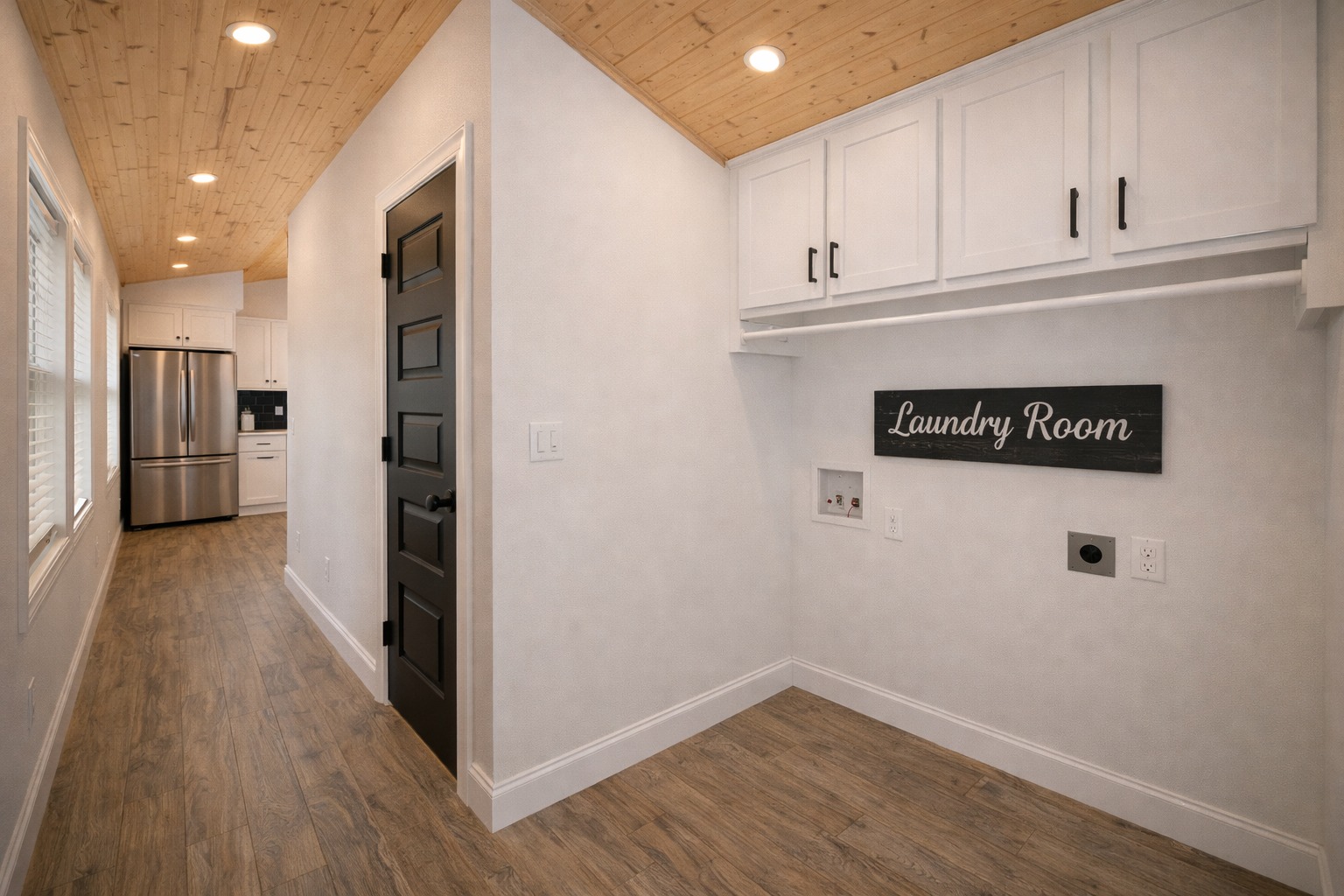 Sleek laundry room with white cabinets, wood ceiling, and flooring. "Laundry Room" sign adds charm. Adjacent to bright, modern kitchen with stainless appliances.