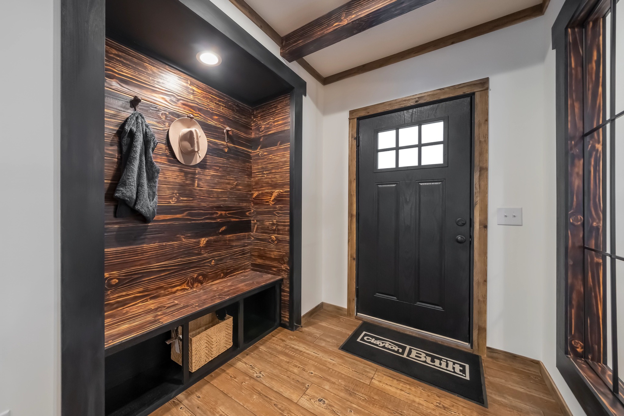 Rustic entryway with a wooden accent wall featuring a coat hook holding a jacket and hat. Black door, bench, and woven basket; log cabin vibe.