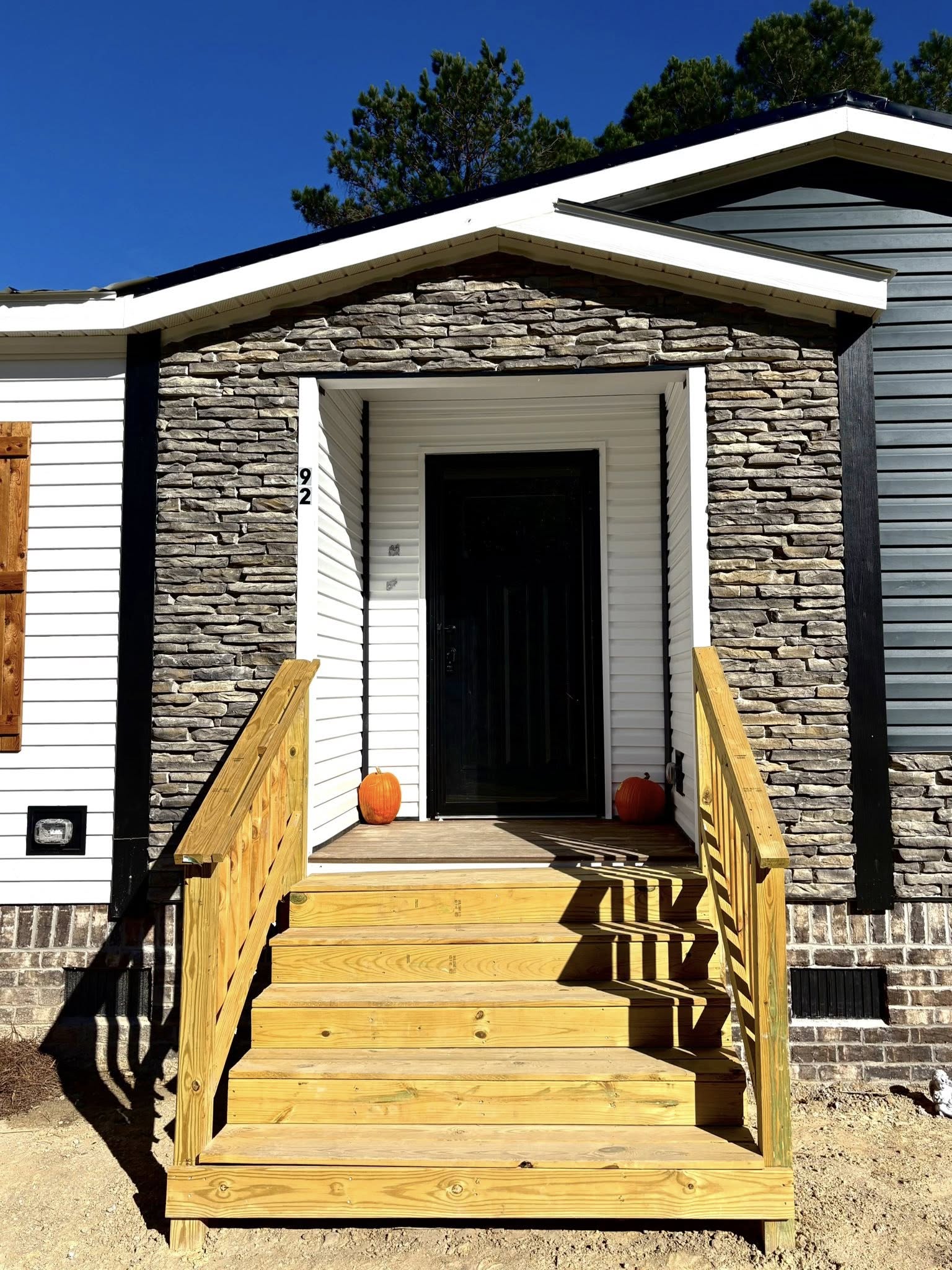 Front entrance of a house with a stone facade, featuring wooden stairs and railings. Two pumpkins sit on either side, creating a welcoming autumnal feel.