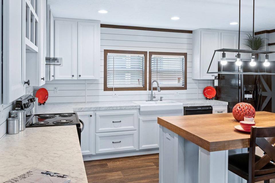 Modern kitchen with white cabinetry, wood countertops, and shiplap walls. A farmhouse sink is beneath two windows. Warm lighting adds a cozy feel.