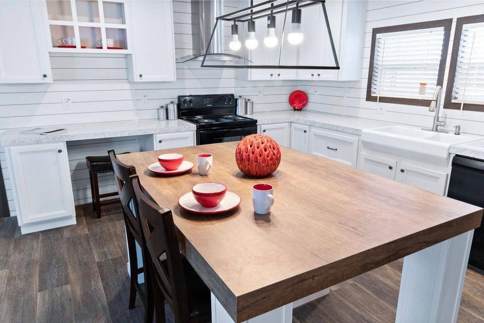 Modern kitchen with white cabinets and wood countertops. A wooden table features red bowls and cups, under a sleek light fixture, conveying warmth.