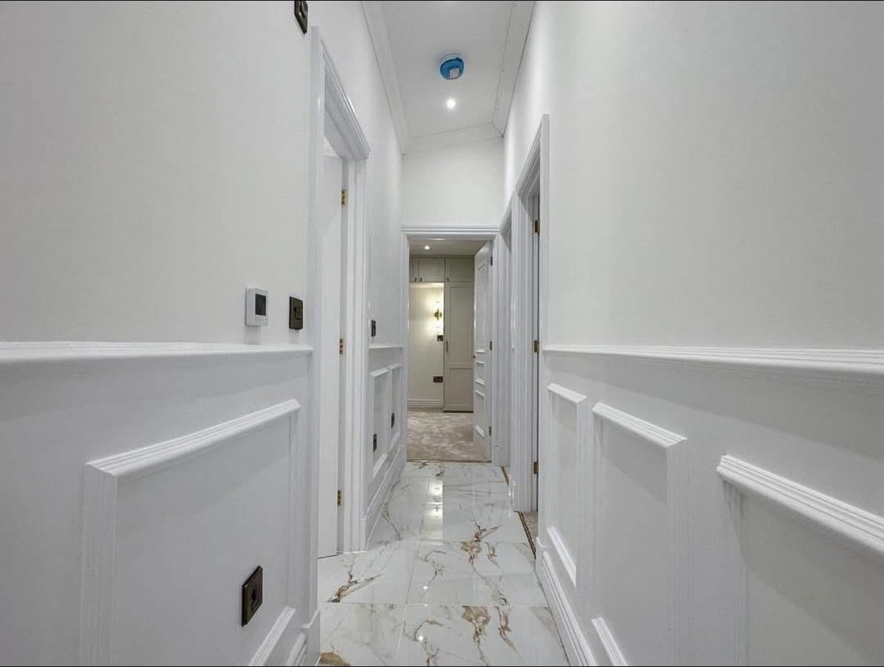 Bright hallway with glossy white walls and marble floor tiles featuring gold veining. Multiple doors lead off the corridor, creating a modern, elegant ambiance.