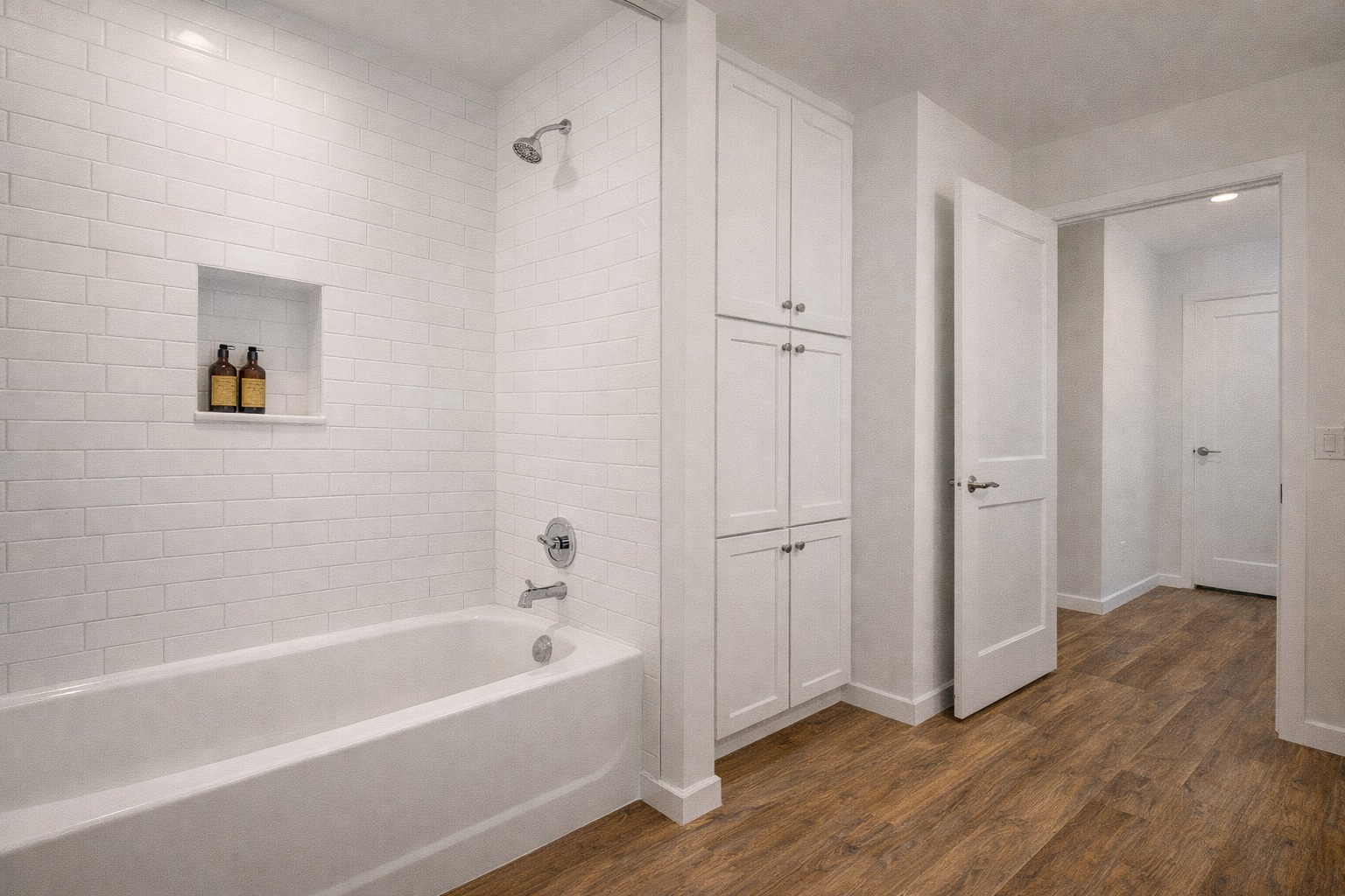 A modern bathroom with white subway tiles surrounding a bathtub. Built-in shelf holds bottles. White cabinets, wood floor, open door to hallway. Minimalist and clean.
