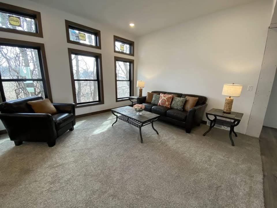 A cozy living room with beige carpet features a dark sofa and matching armchair, accent pillows, two lamps, and a glass coffee table. Large windows let in natural light.