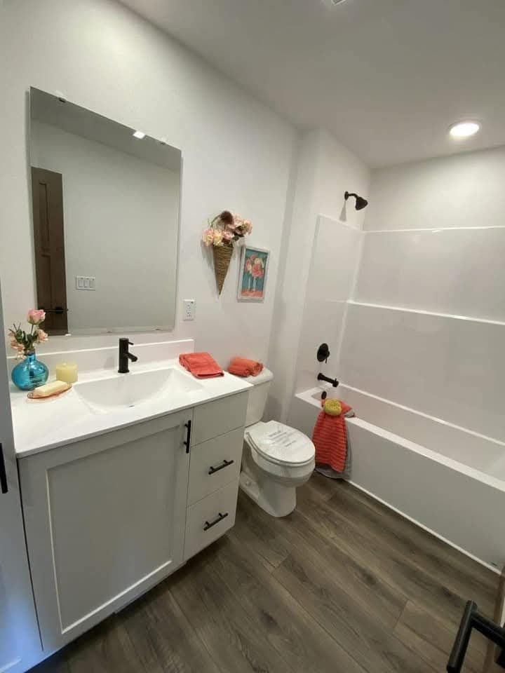 A modern bathroom with wood flooring, featuring a white vanity with a black faucet, colorful towels, floral decor, and a shower-tub combo. Bright and inviting.