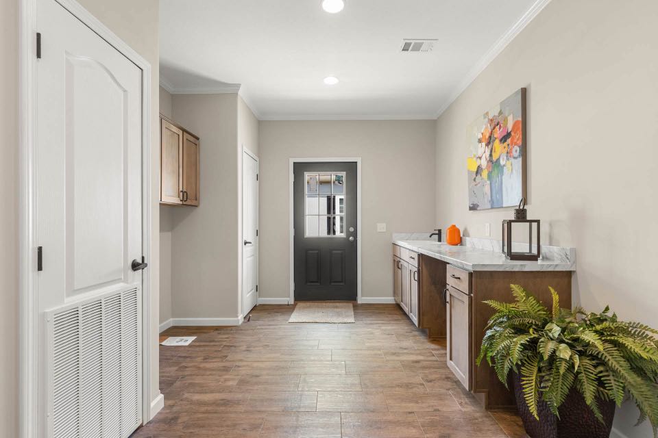 Bright mudroom with wood flooring, white walls, and a dark door. A countertop holds a colorful flower painting and a fern in the corner adds greenery.