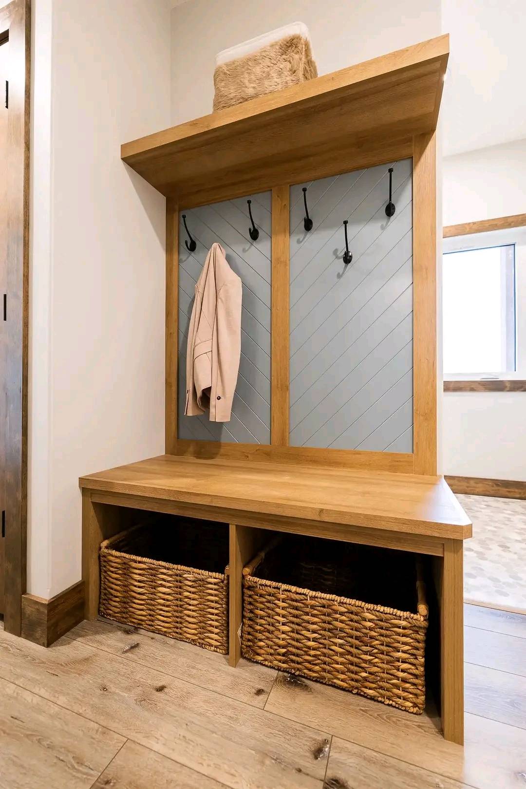 Wooden entryway bench with hooks, a beige coat hung, and wicker baskets beneath. Shelf above holds a textured basket. Bright and tidy atmosphere.