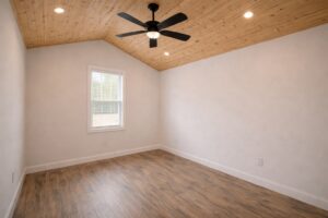 Bright empty room with a wooden ceiling and floor, white walls, a ceiling fan, and a window with blinds. The atmosphere is clean and modern.