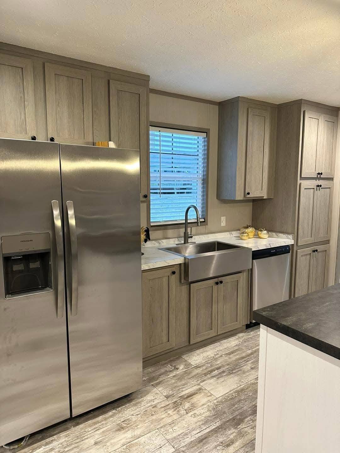 Modern kitchen with stainless steel fridge, farmhouse sink, and wooden cabinets. A window above the sink and gray tile floors add a warm, inviting feel.