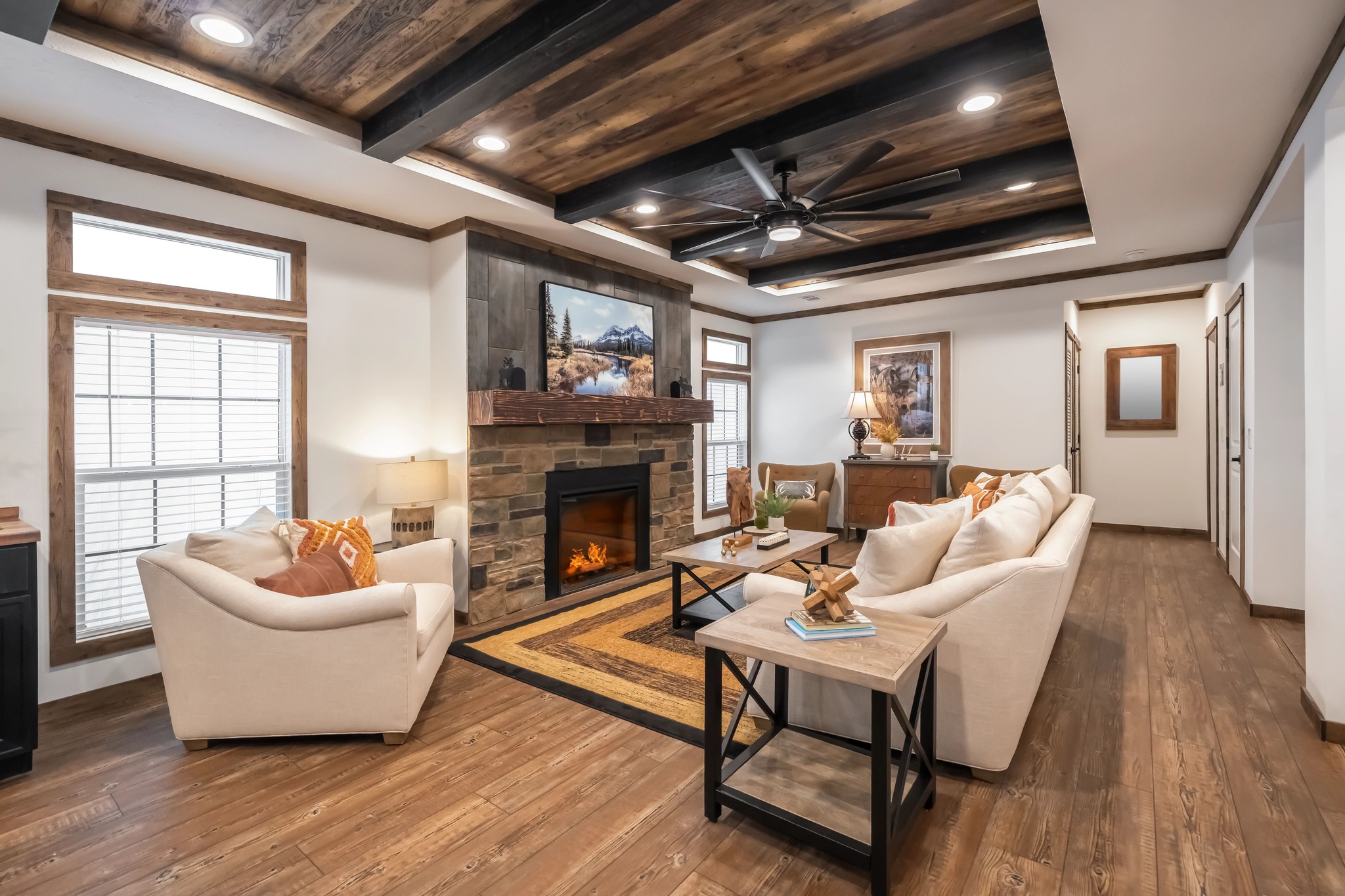 Warm and inviting living room with wood accents features beige sofas and armchairs around a stone fireplace. Large windows and rustic decor create a cozy ambiance.