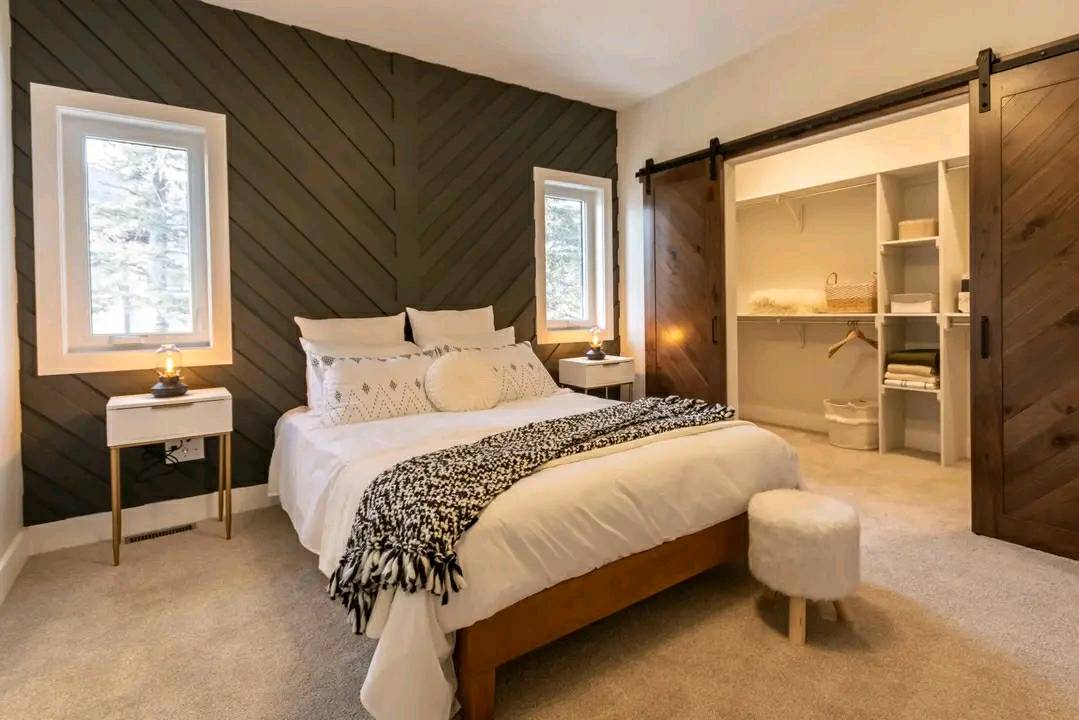 A cozy bedroom with a stylish black accent wall and chevron pattern. Features a plush bed with decorative pillows, flanked by two small lamps. An open closet with wooden sliding doors is partially visible. Soft, neutral tones create a warm, inviting atmosphere.