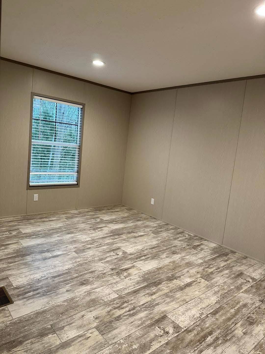 Empty room with beige walls and wood-patterned vinyl flooring. A window with closed blinds is on the left wall, and the room is well-lit with ceiling lights.