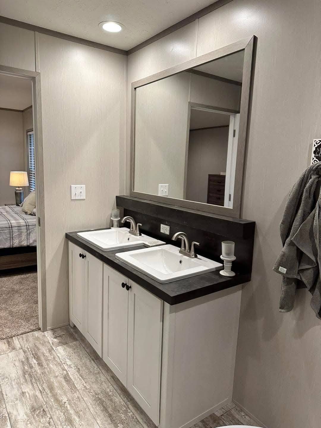 Modern bathroom with a double sink vanity featuring a large mirror. White cabinets and sleek fixtures set a clean, elegant tone. Cozy bedroom visible in the background.