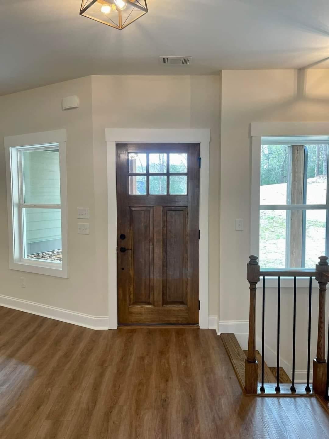 Modern entryway with wooden door and glass panes, flanked by two large windows. Warm wood flooring and stairs enhance the inviting, cozy ambience.