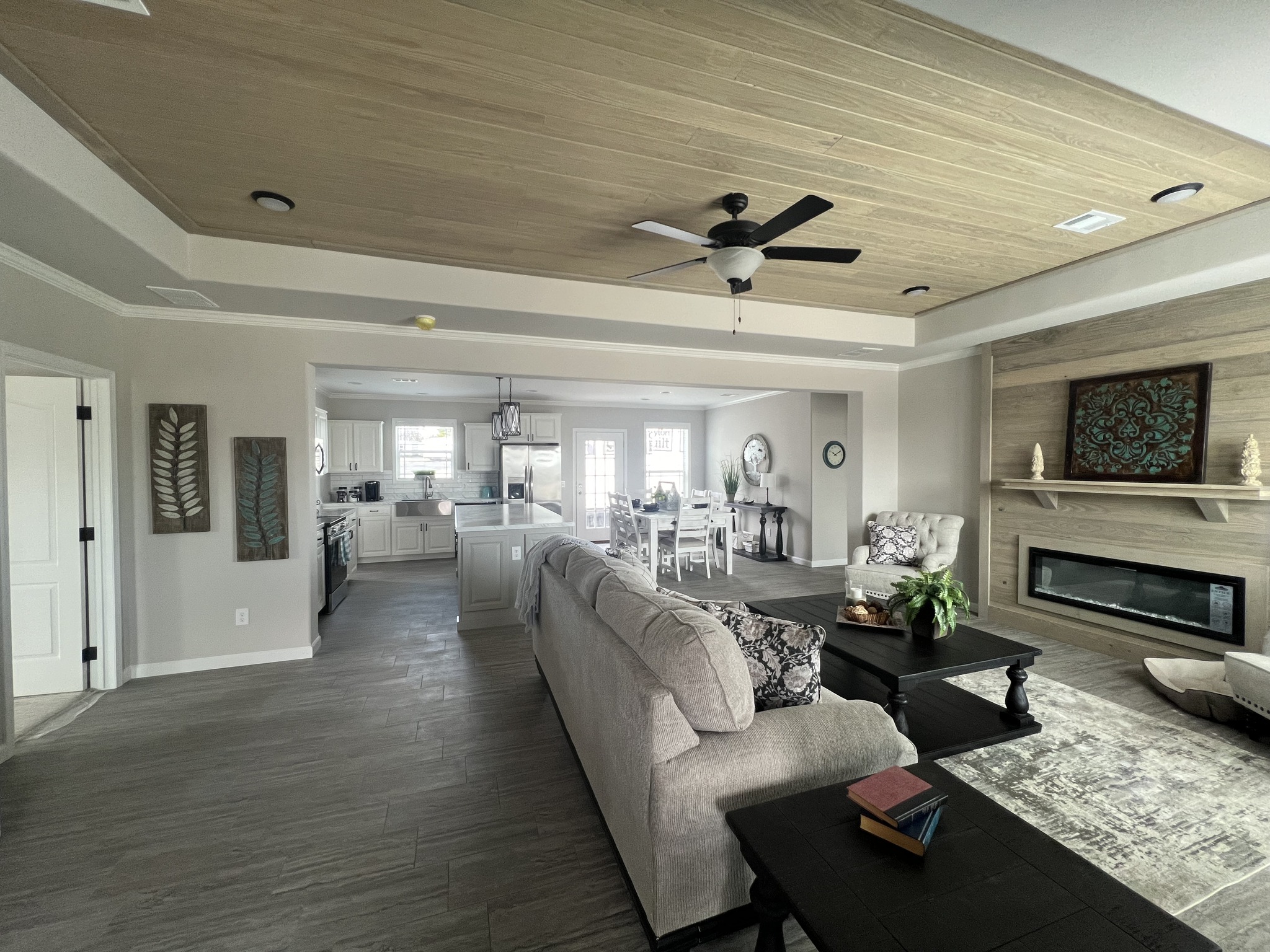 Spacious living room with beige sofa and black coffee table, leading to a bright kitchen and dining area. Ceiling fan, wall art, and a modern fireplace add elegance.