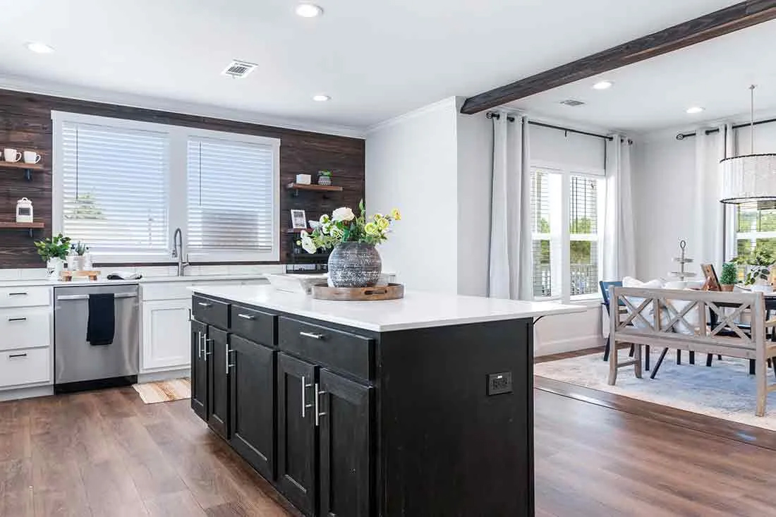 Modern open kitchen and dining area with wood floors. Features a white island, dark cabinets, large windows, and a stylish dining table, creating a bright, welcoming ambiance.