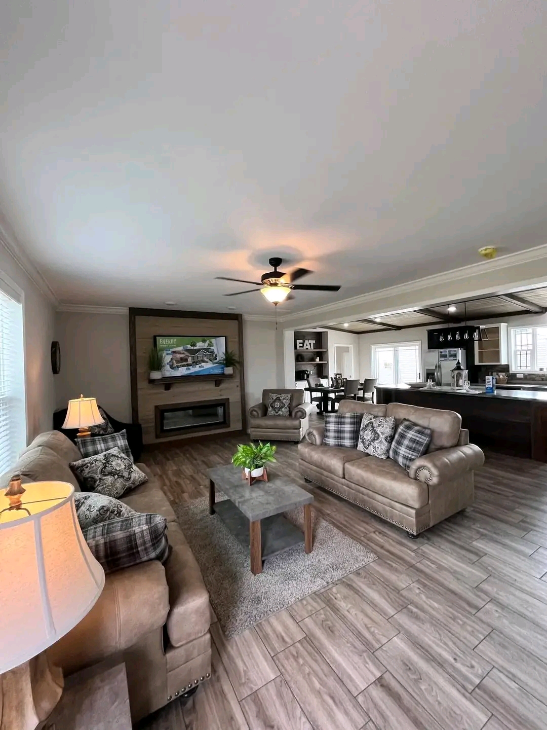 Spacious living room with cozy beige sofas and checkered cushions. A ceiling fan, large TV, and fireplace enhance the warm, inviting ambiance. Open layout to kitchen.