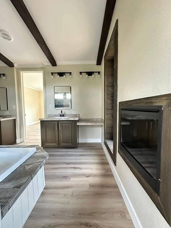 Spacious bathroom with a modern, cozy feel. Features a bathtub, dual mirrors with lights, wooden cabinets, and a wall-mounted fireplace. Light wood flooring.