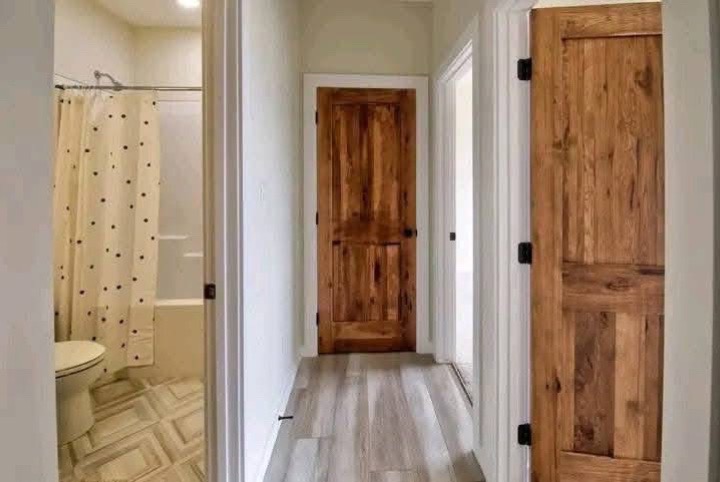 A hallway with light wood flooring features three wooden doors. The left door leads to a bathroom with a beige polka dot shower curtain and patterned tile floor.