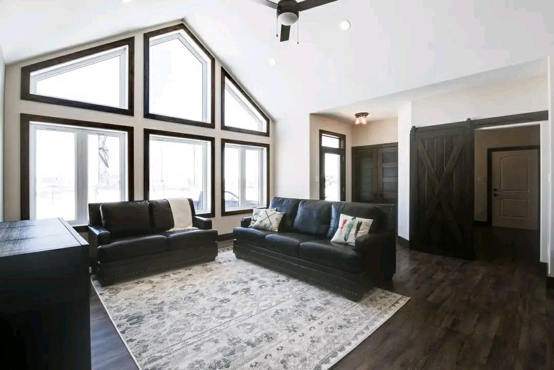 A spacious living room with high triangular windows allowing ample light. It features two black leather sofas on a patterned rug, dark wood floors, and a sliding barn door, creating a cozy and modern feel.