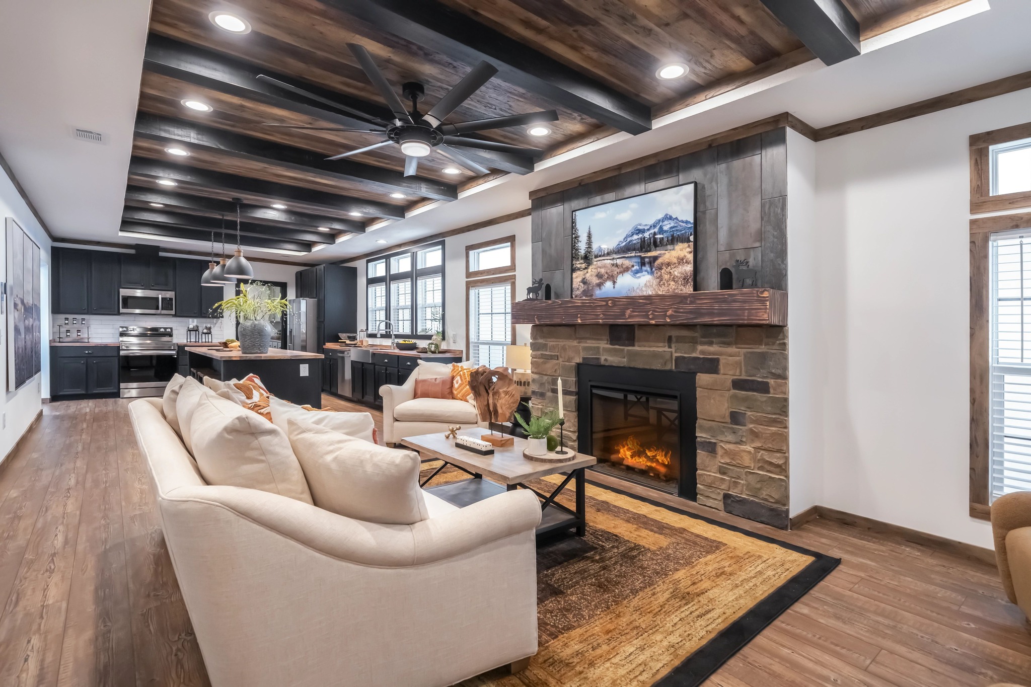 Stylish open-concept living room and kitchen with wood beam ceiling. Features a stone fireplace, cream sofas, rustic accents, and modern appliances.